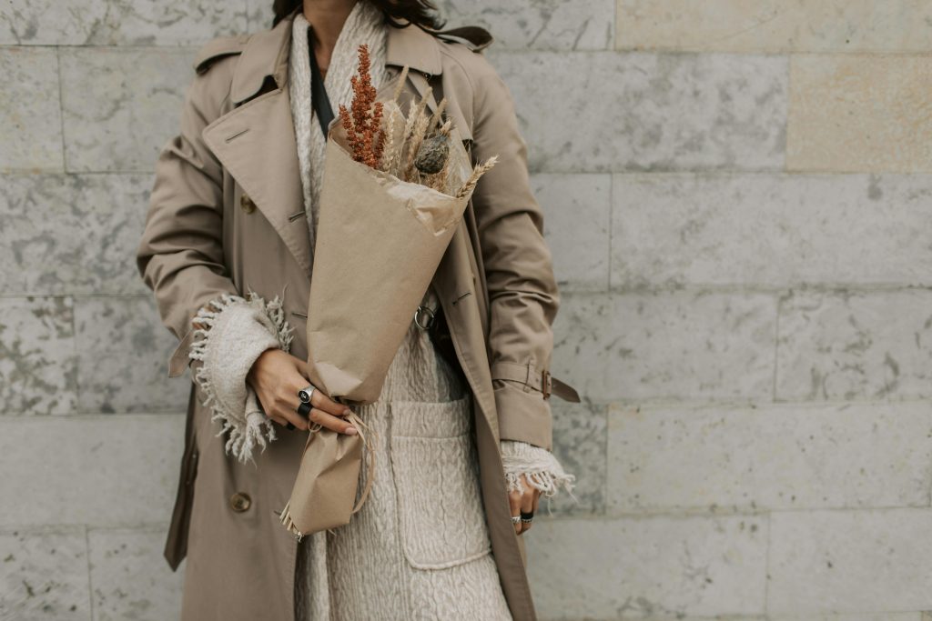 woman standing against a grey brick wall wearing a beige trench coat and holding dried flowers
